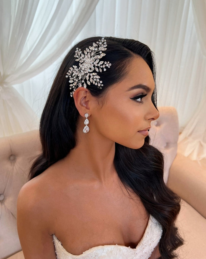 A bride in a strapless gown sits on a beige sofa against sheer white curtains. Her dark hair is styled with the Elegant Bridal Comb, style no. 242512, and her dangling Cubic Zirconia and Swarovski earrings complement her soft expression as she gazes to the side.