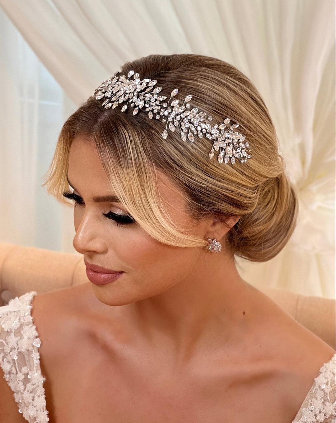 A woman with neatly styled blonde hair in a low bun, accented by the Elegant Crystal Cluster Wedding Hair Vine style no. 242517. She wears a lace bridal gown and star-shaped earrings against a softly draped fabric background.
