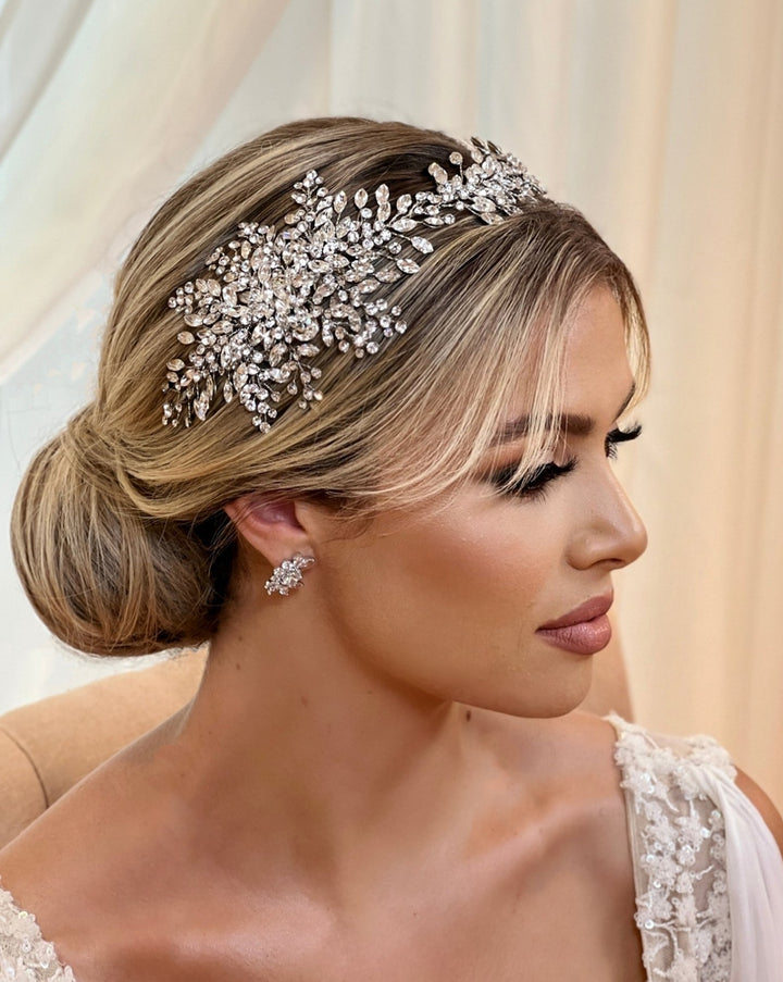 A person with blonde hair in a bun wears an ornate Swarovski crystal headpiece and matching CZ bridal earrings. They are dressed in a bridal gown, looking forward, against a soft focus curtain backdrop.