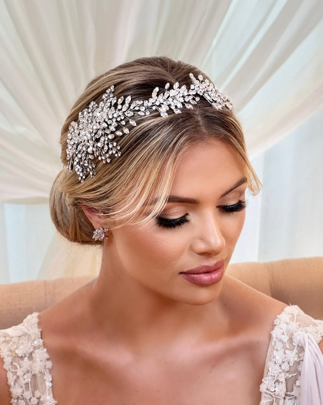 A woman with blond hair elegantly styled in an updo dons the Asymmetrical Cluster Crystal Hair Vine style no. 242641 . Her soft makeup and serene expression complement the delicate, lacy gown against a curtain backdrop, creating an ethereal bridal look.