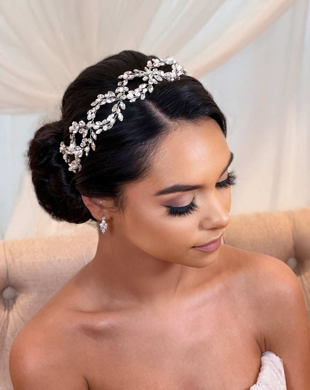 A dark-haired woman styled in an updo with a Classic Pearl and Crystal Looping Swarovski Hair Vine sits on a beige chair, wearing subtle makeup and a strapless gown. She gazes down gently, with a white curtain behind her enhancing her elegant bridal look.