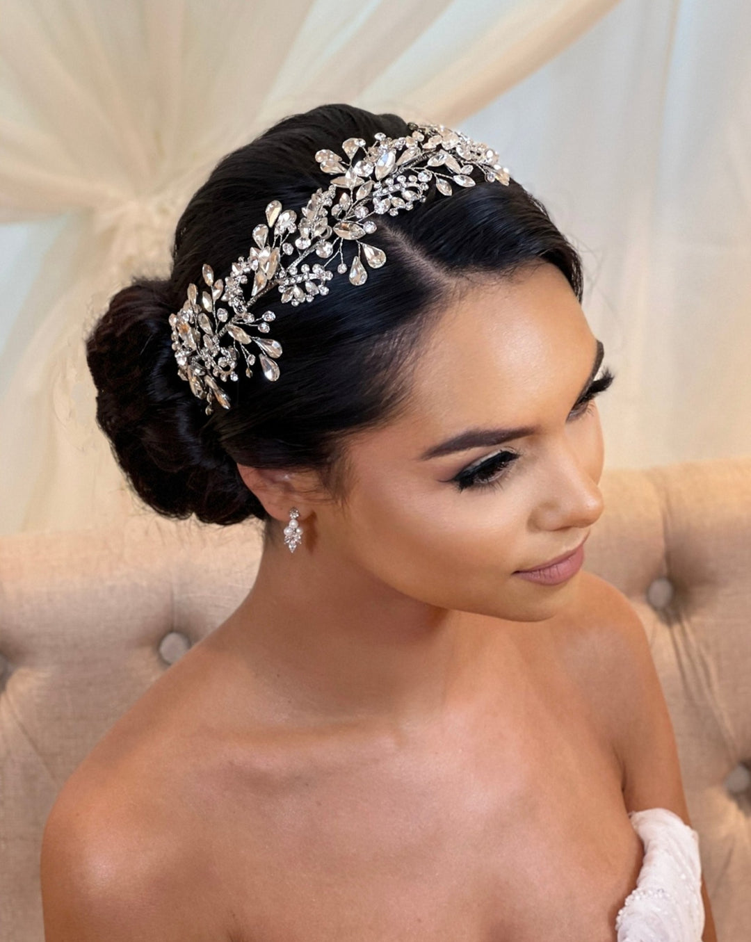 A woman with dark hair in an elegant bun wears the Teardrop Swarovski Crystal Vine tiara (style no. 242696), showcasing leaf and floral designs. She dons a white strapless gown and sits on a tufted beige chair, gazing gracefully to the side.