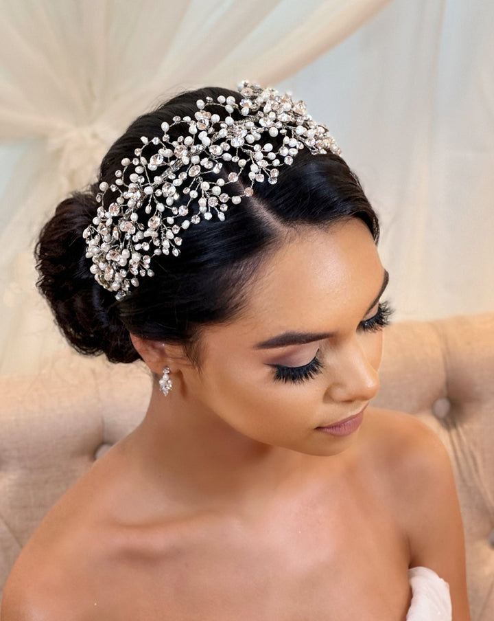 A woman with dark hair styled in an elegant bun wears the Classy Bridal Headband style no. 242676 and matching earrings. Her eyes are closed as she sits on a beige chair against a soft background, creating a serene and elegant atmosphere.