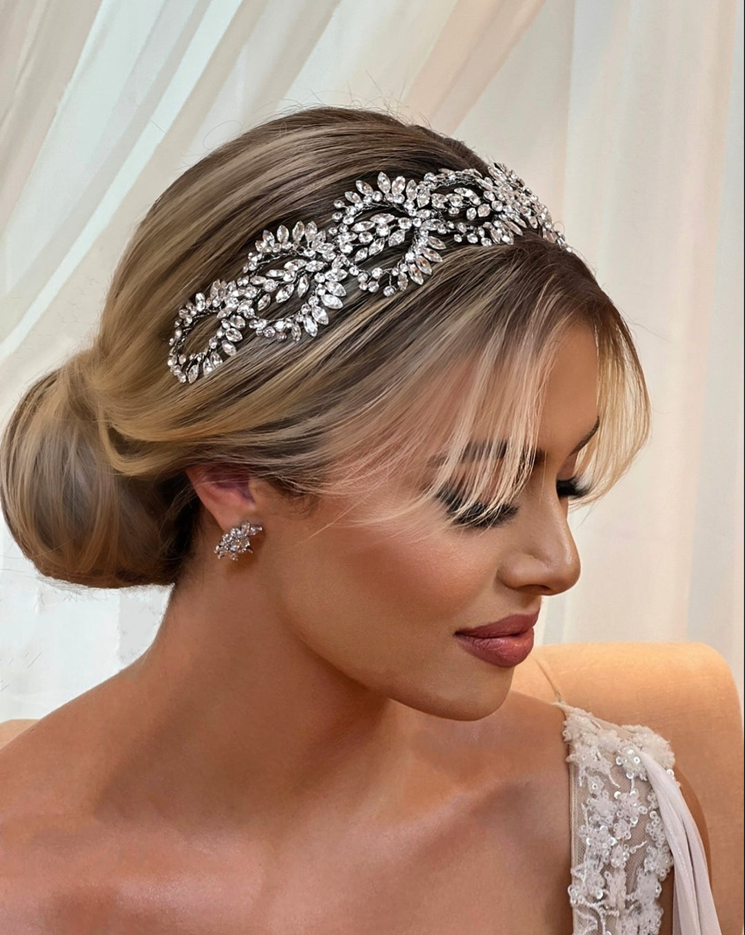 A woman with blonde hair in a low bun wears the Crystal Wreath Vine, style no. 242643, adorned with matching earrings. She gazes downward, surrounded by soft lighting and sheer curtains.