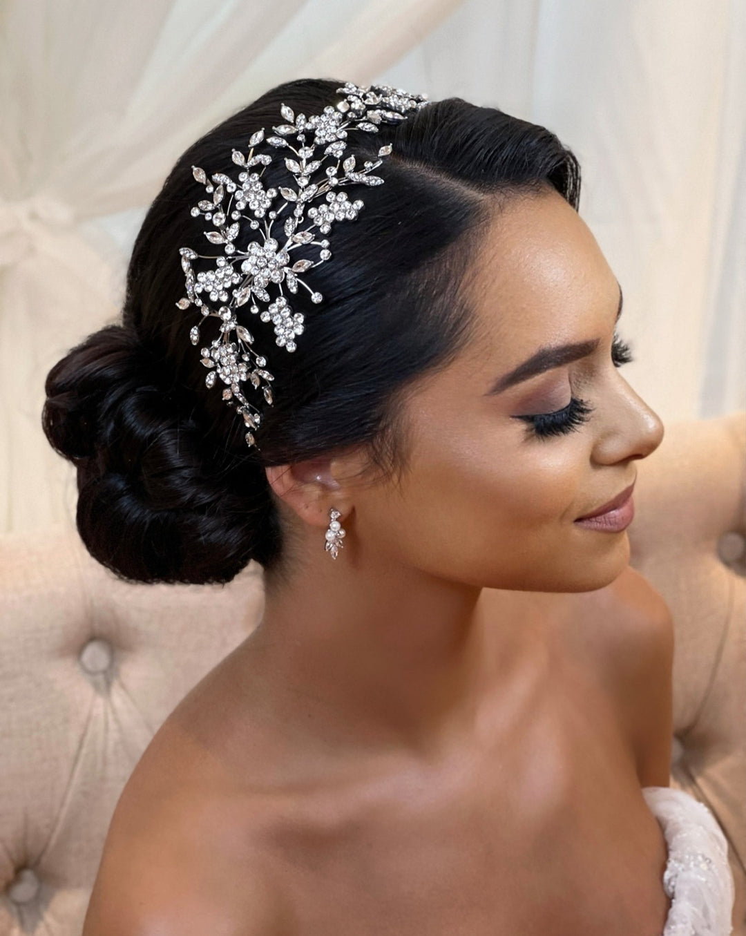 A woman with dark hair in an elegant updo wears the Asymmetrical Crystal Flower Headband style no. 242729 and pearl and diamond drop earrings. Her eyes are closed, and she smiles softly against a tufted, beige backdrop.