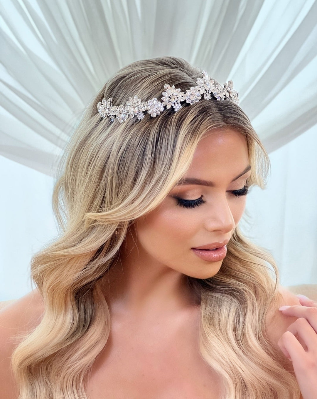 A person with wavy blonde hair wears a silver Floral Crystal Bridal Tiara style no. 80057, as they gaze downward under draped white fabric for a soft, refined look.