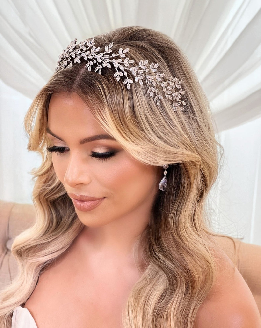 A woman with long blond hair wears a sparkling Swarovski Romantic Crystal Headband (style no. 7930116) and delicate earrings. Seated against a draped light background, she gazes down serenely.