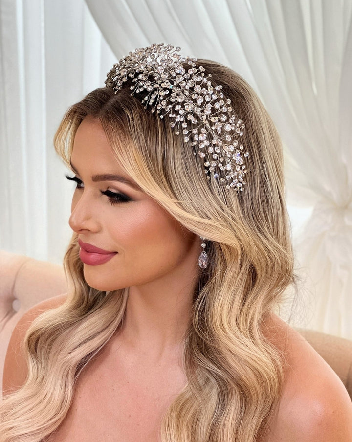 A woman with long, wavy blonde hair and soft makeup wears a sparkly Bursting Crystal Headband style no. 3910361 resembling a Swarovski wedding crown, and matching dangling earrings. She is seated against a light, draped background, looking to the side with a calm expression.