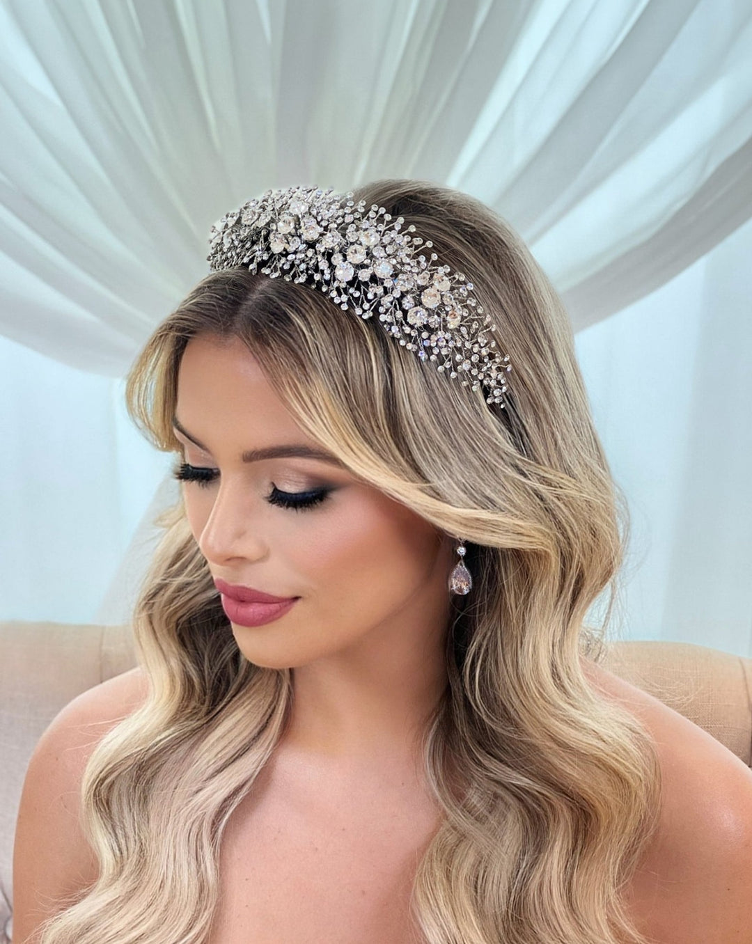 A person with long, wavy blond hair wears the Bold and Beautiful Crystal Bridal Headband style no. 391022, featuring ornate details and Swarovski crystals. They have soft makeup and pink lipstick, gazing downward against sheer white curtains.