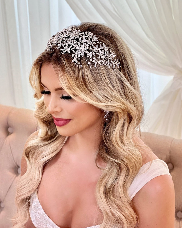 A woman with long blonde hair styled in waves sits on a beige tufted chair, wearing a Wide Floral Crystal Headband style no. 242689 and a white off-shoulder dress. The background is softly draped with white fabric.