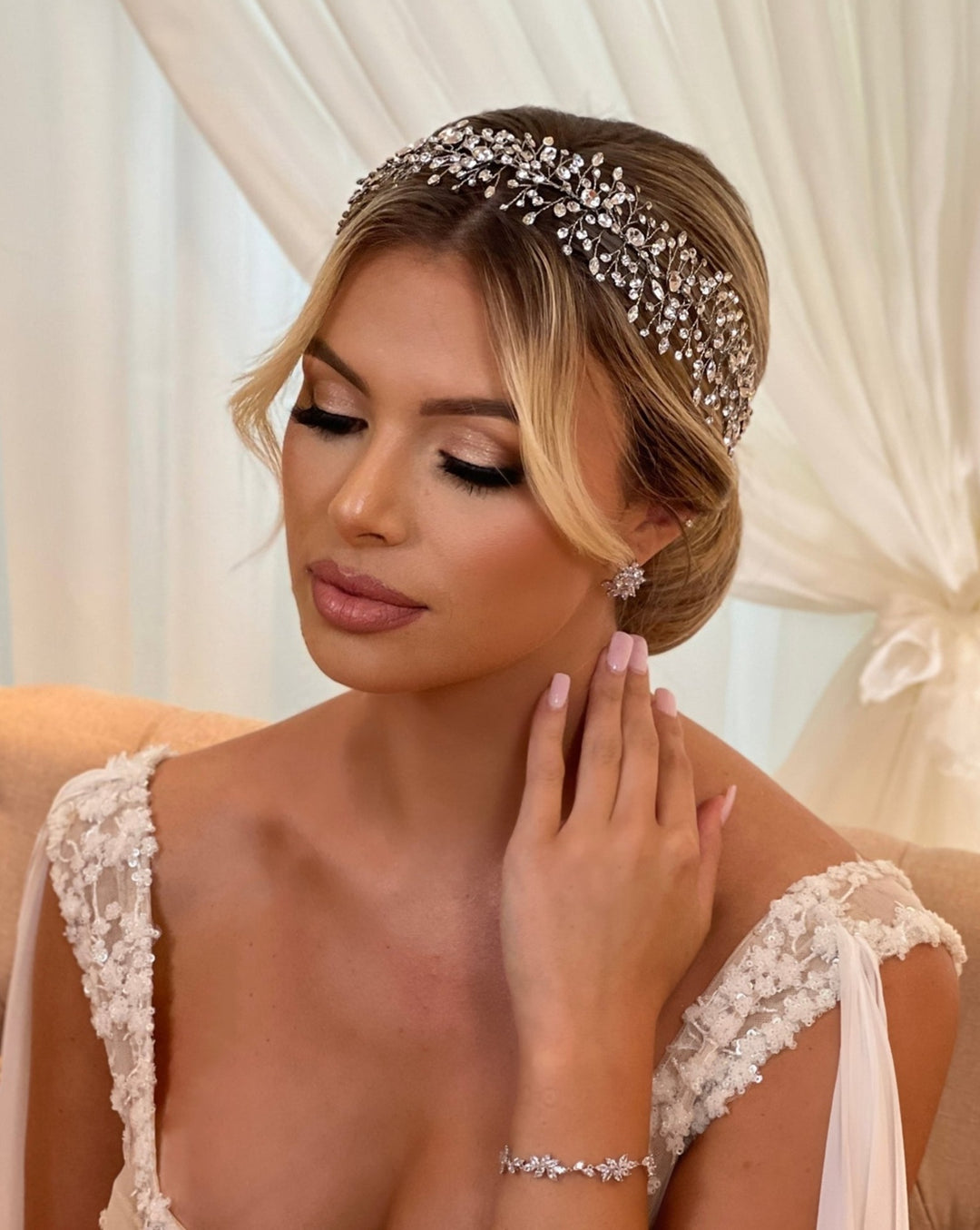 A woman in a white lace gown wears the Elegant Branching Swarovski Crystal Hair Vine (style no. 242699)  with matching wedding jewelry, including earrings and a bracelet. Her hair is styled in an updo with elegant makeup, seated against a light curtain backdrop.