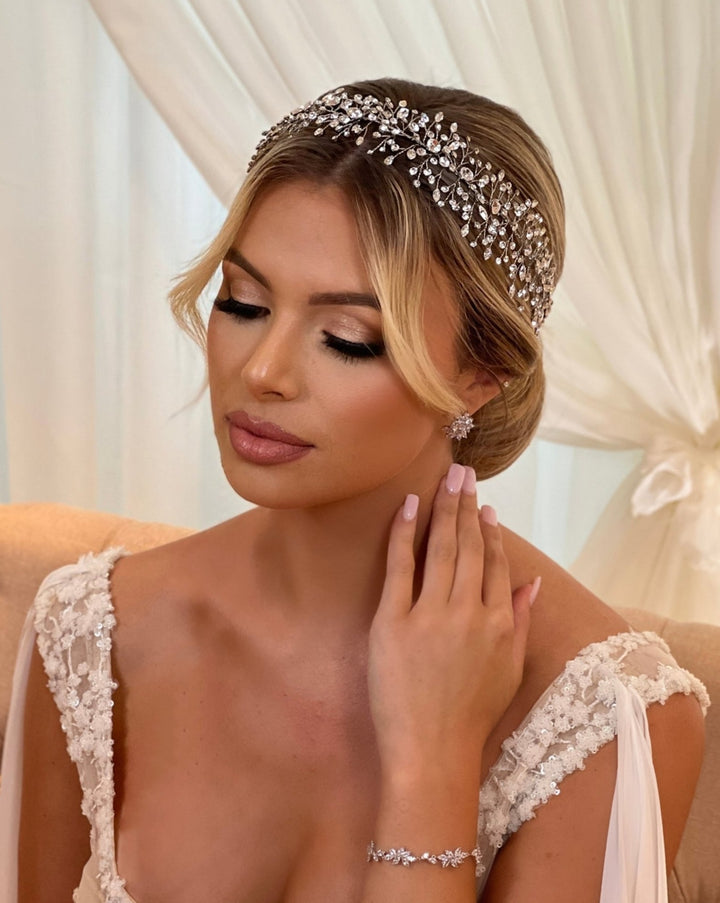 A woman in a white lace gown wears the Elegant Branching Swarovski Crystal Hair Vine (style no. 242699)  with matching wedding jewelry, including earrings and a bracelet. Her hair is styled in an updo with elegant makeup, seated against a light curtain backdrop.