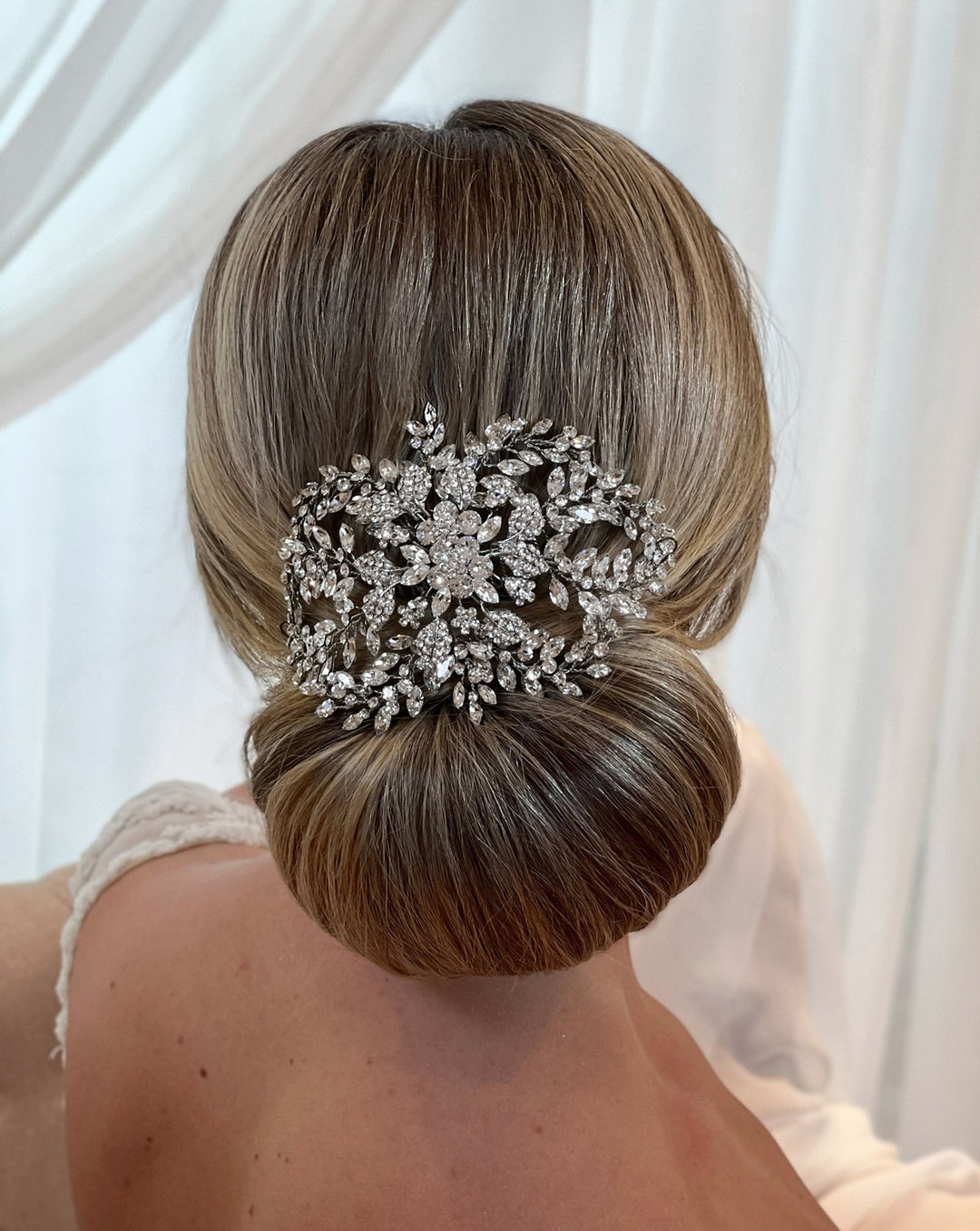 A woman wears a white lace dress, her hair styled in a sleek bun adorned with the  Bloom Swarovski Crystal Hair Comb, style no. 242717 , showcasing intricate floral and leaf designs against softly draped white fabric.