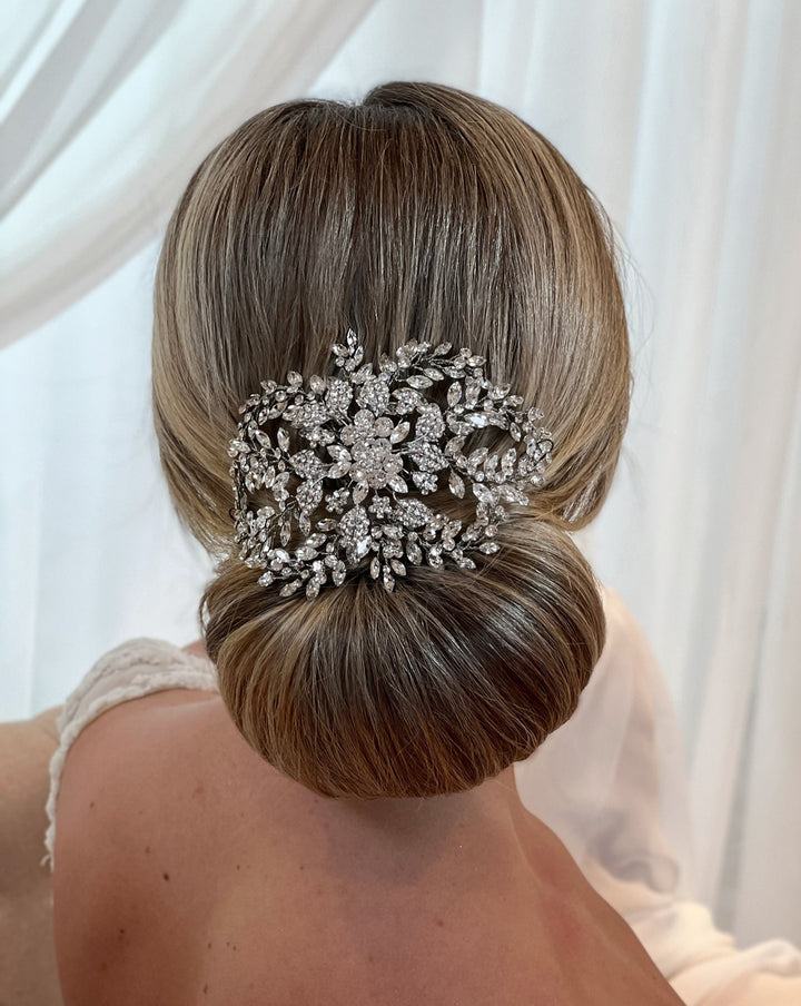 A woman wears a white lace dress, her hair styled in a sleek bun adorned with the  Bloom Swarovski Crystal Hair Comb, style no. 242717 , showcasing intricate floral and leaf designs against softly draped white fabric.