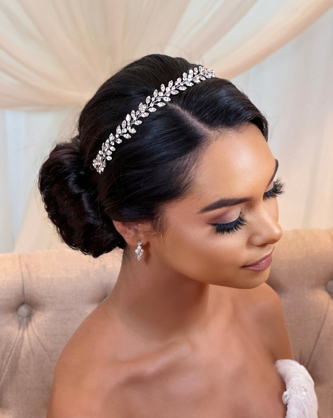 A woman with a side profile view wears the sparkling Traditional Swarovski  Crystal Vine (style no. 242710) wedding crown and elegant CZ bridal earrings. Her dark hair is styled in a low bun, she has long eyelashes, wears subtle makeup, and stands against a softly draped background exuding elegance.