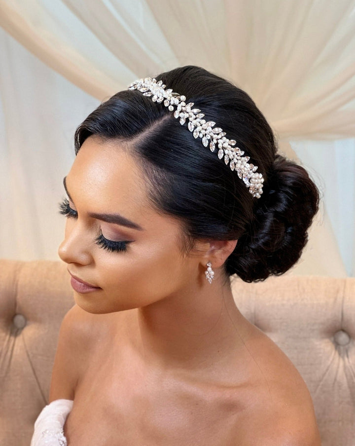 A woman with styled black hair in an elegant updo adorned with the Traditional Swarovski Crystal Hair Vine headband style no. 242710 wears dangling CZ wedding earrings. She sits against a light-colored tufted backdrop, wrapped in soft white fabric.