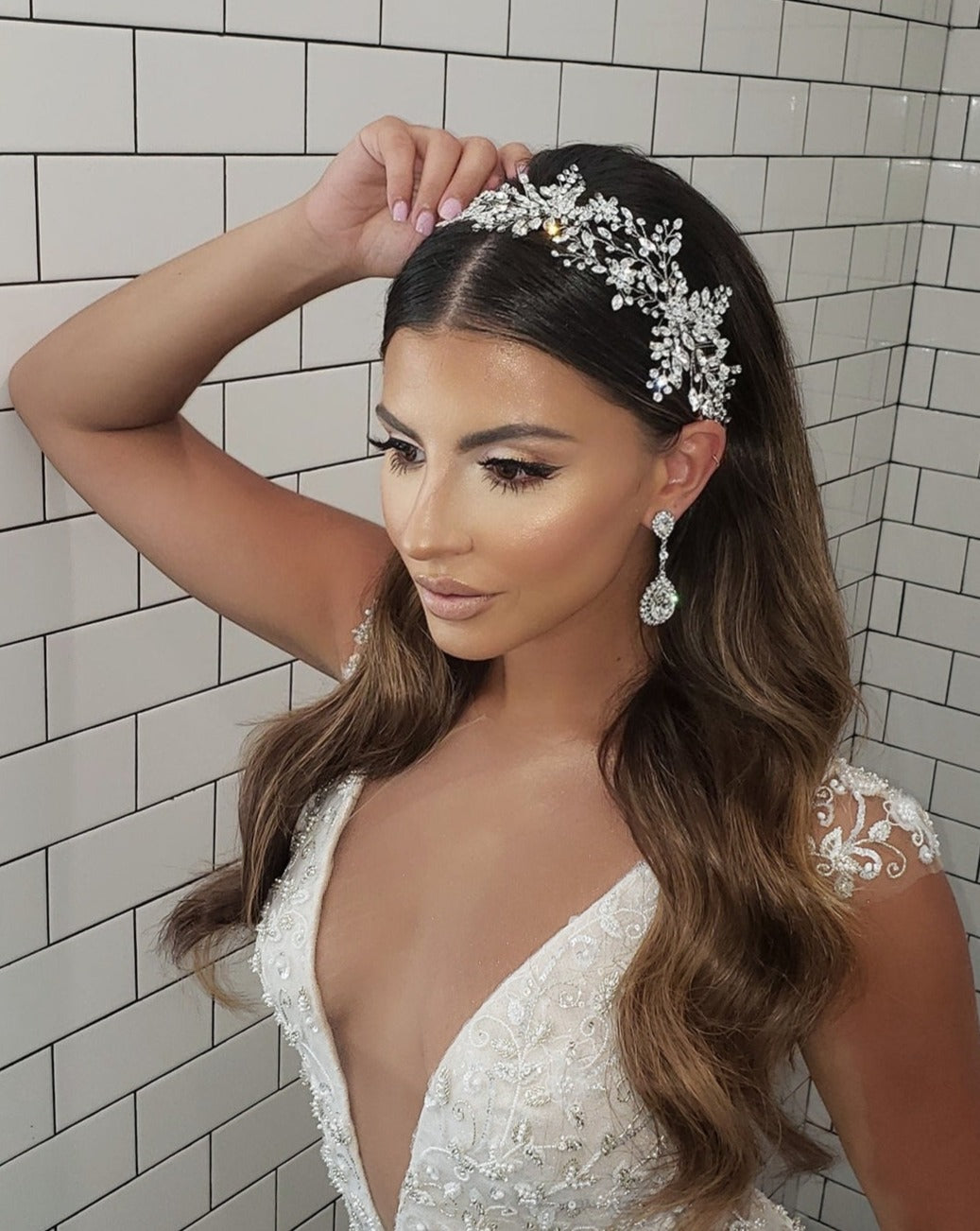 A woman with long, wavy hair accents her hair with the Stunning Swarovski Crystal Bridal Hair Vine style no. 242594 poses against a tiled wall in an ornate white dress featuring lace details and large dangling earrings, exuding elegance and grace.