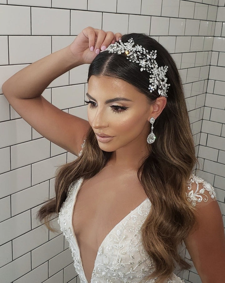 A woman with long, wavy hair accents her hair with the Stunning Swarovski Crystal Bridal Hair Vine style no. 242594 poses against a tiled wall in an ornate white dress featuring lace details and large dangling earrings, exuding elegance and grace.