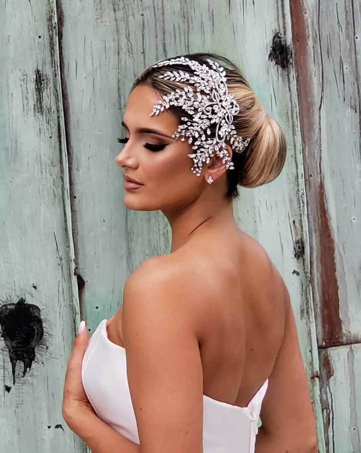 A woman in a strapless white dress stands with her back and side to the camera, wearing a Glamorous Crystal Hair Comb style no. 242535. Her hair is styled in a bun against a backdrop of weathered wooden panels.
