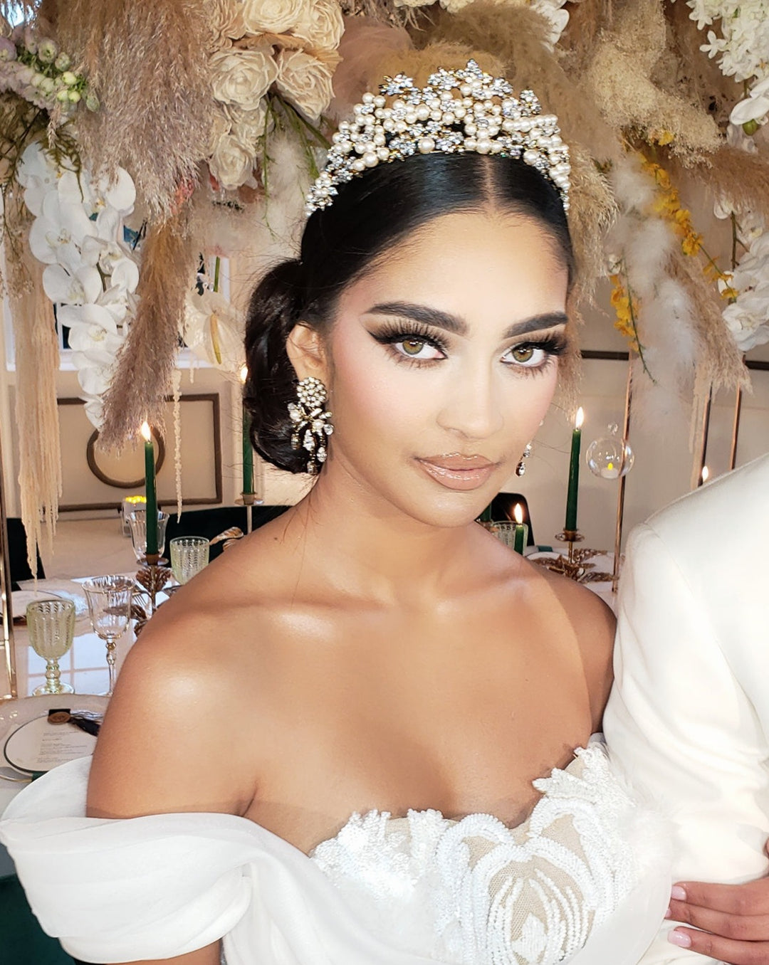 A woman in a white dress smiles softly, adorned with a Swarovski crystal floral headpiece and elegant makeup highlighted by long eyelashes. A Pearl and Crystal Bridal Tiara (style no. 80044) twinkles amid her curls, while behind her, a flower-adorned table with candles and glassware sets an elegant festive scene.