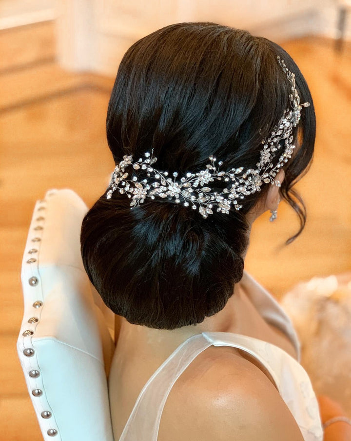 A person with a stylish updo wearing the Delicate Weaving Swarovski  Crystal Vine (style no. 242465) i, sitting on a white chair with studded details, against a softly blurred background.