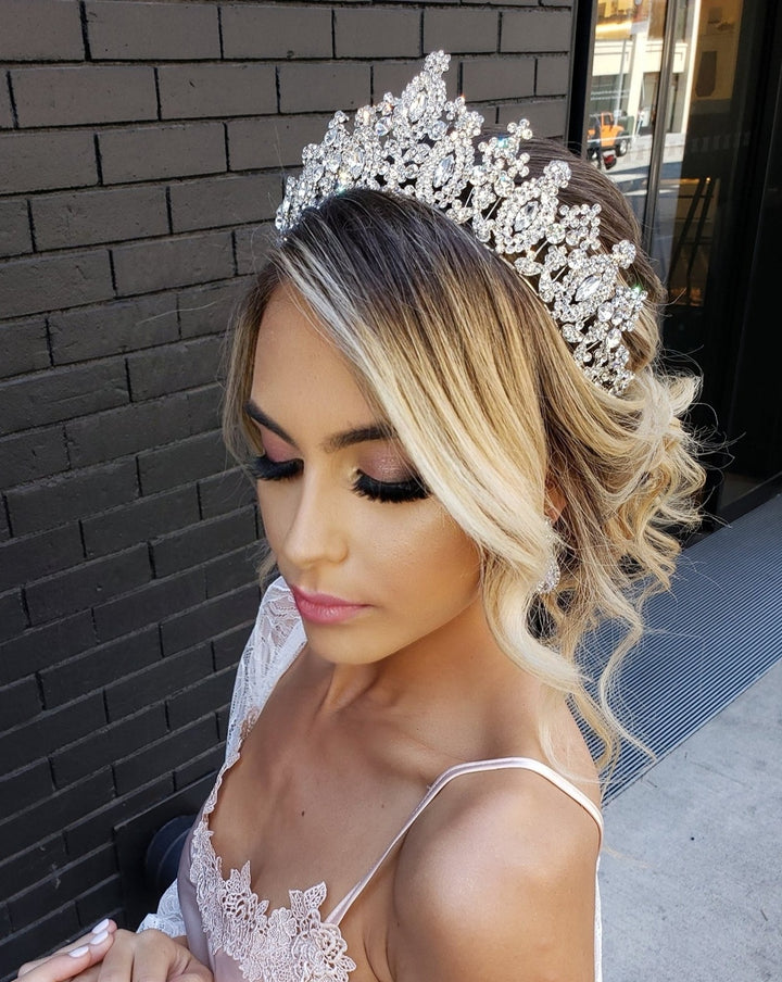 A woman in a light pink lace dress and a Swarovski crystal-encrusted Royal Wedding Tiara style no. 80034 stands against a dark brick wall, her hair elegantly styled in a loose updo. She looks down with eyes closed, highlighting her eye makeup.
