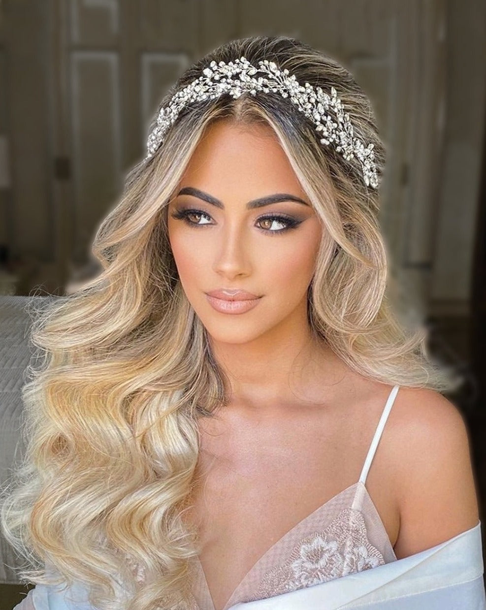 A woman with long, wavy blonde hair wears the Delicate Weaving Swarovski Crystal Vine style no. 242465 as a headband. Her defined makeup enhances the elegance of her white, lace-trimmed garment against a softly blurred background.