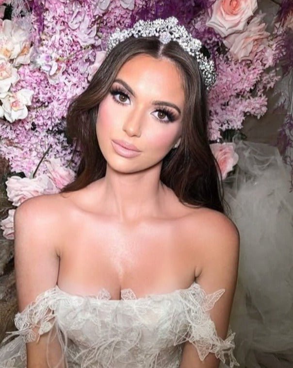 A bride in a strapless lace wedding dress, wearing the Swarovski Crystal Wedding Tiara style no. 80039, poses before pink and purple flowers. Her long brown hair flows in loose waves, complemented by soft, elegant makeup.