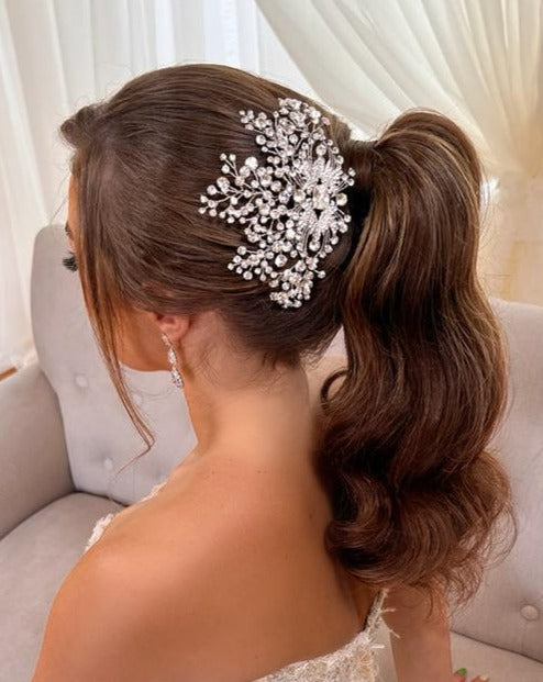 Adorned with the intricate Art Deco Crystal Comb style no. 242693, a woman with long, wavy brown hair styled in a ponytail complements her elegant chandelier earrings and delicate Swarovski hair vine while sitting on a light-colored sofa framed by sheer curtains.