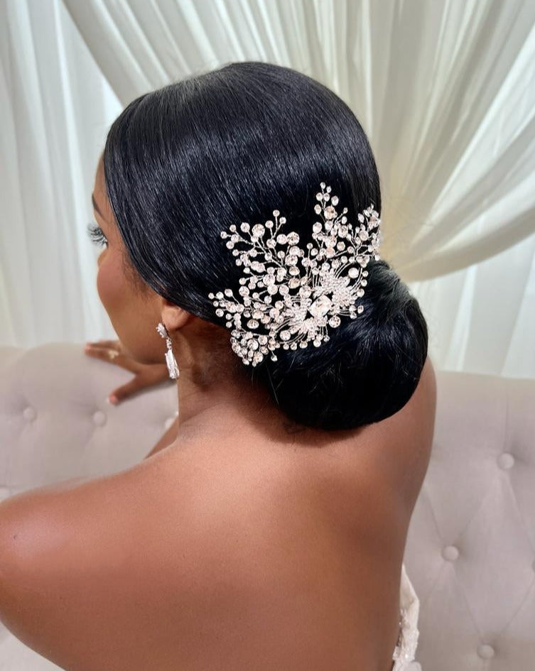 A woman with sleek black hair in a low bun faces away, hair hairstyle adorned by the Art Deco Crystal Bridal Comb style no. 242693. Elegant earrings enhance her look against a soft, light-colored fabric background.