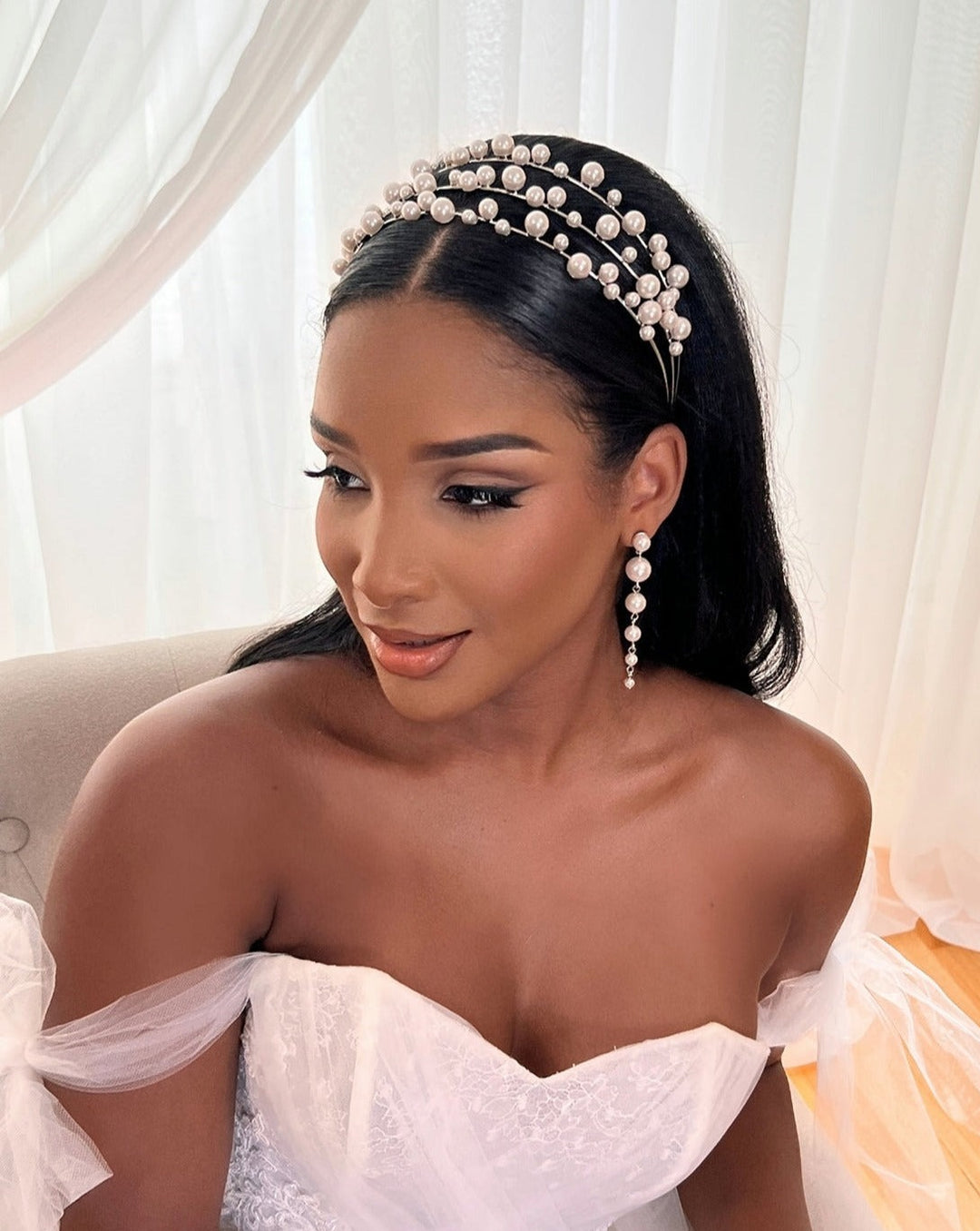 A woman in a white off-shoulder wedding dress with sheer sleeves sits on a chair, wearing the Tiered Pearl Bridal Headband (Style no. 80096) and dangling pearl earrings. Her long dark hair is styled loosely with a Swarovski hair vine. Curtains and soft lighting provide an elegant backdrop.