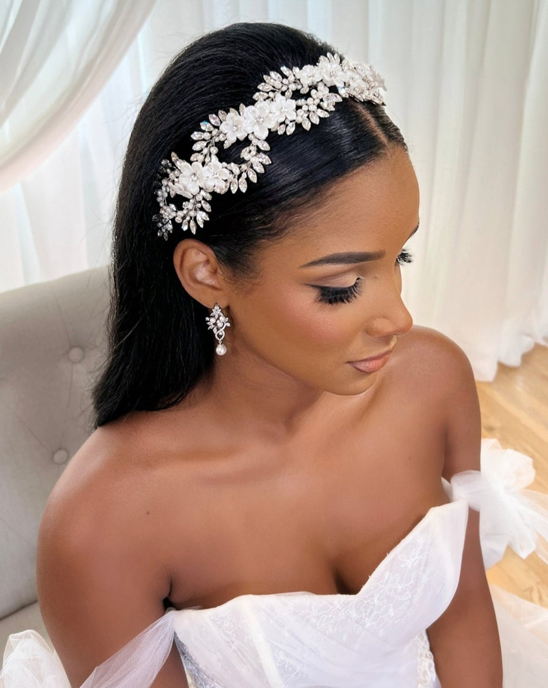 A bride with long black hair wears a Swarovski wedding crown and pearl earrings, showcasing her off-the-shoulder white lace gown. She is seated in a chair against a bright, softly lit background, adorned with the Elegant Looping Flower Vine style no. 242727.