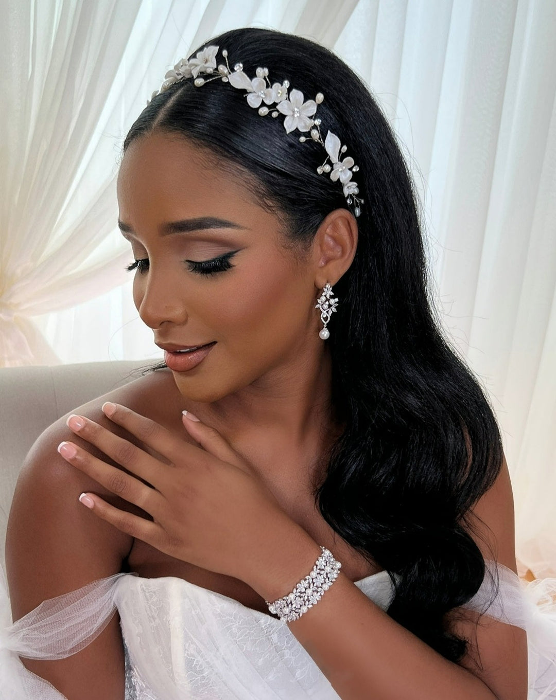 A woman with long dark hair, wearing a Delicate Porcelain Flower and Swarovski Crystal Hair Vine (style no. 242732), elegant earrings, and a bracelet, is dressed in a white off-shoulder gown. She poses gracefully with one hand on her shoulder in front of sheer curtains.