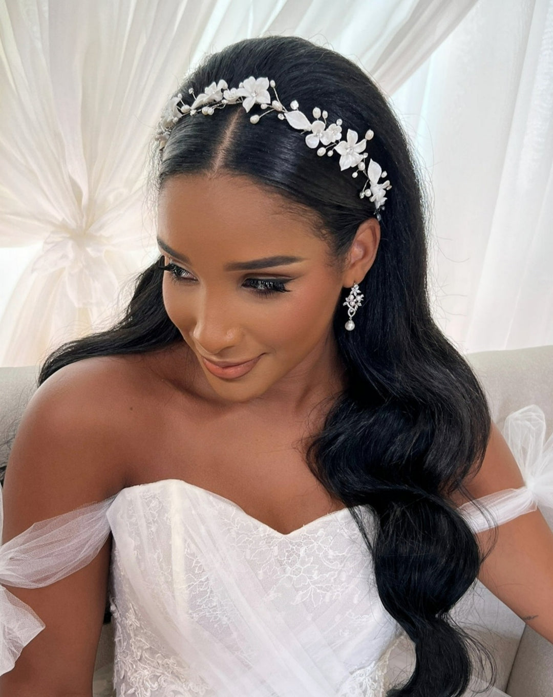 A bride with long, flowing dark hair adorned with the Delicate Porcelain Flower Bridal Hair Vine sits in a white lace wedding dress. The off-shoulder gown features sheer fabric details. She has elaborate eye makeup and elegant floral earrings.