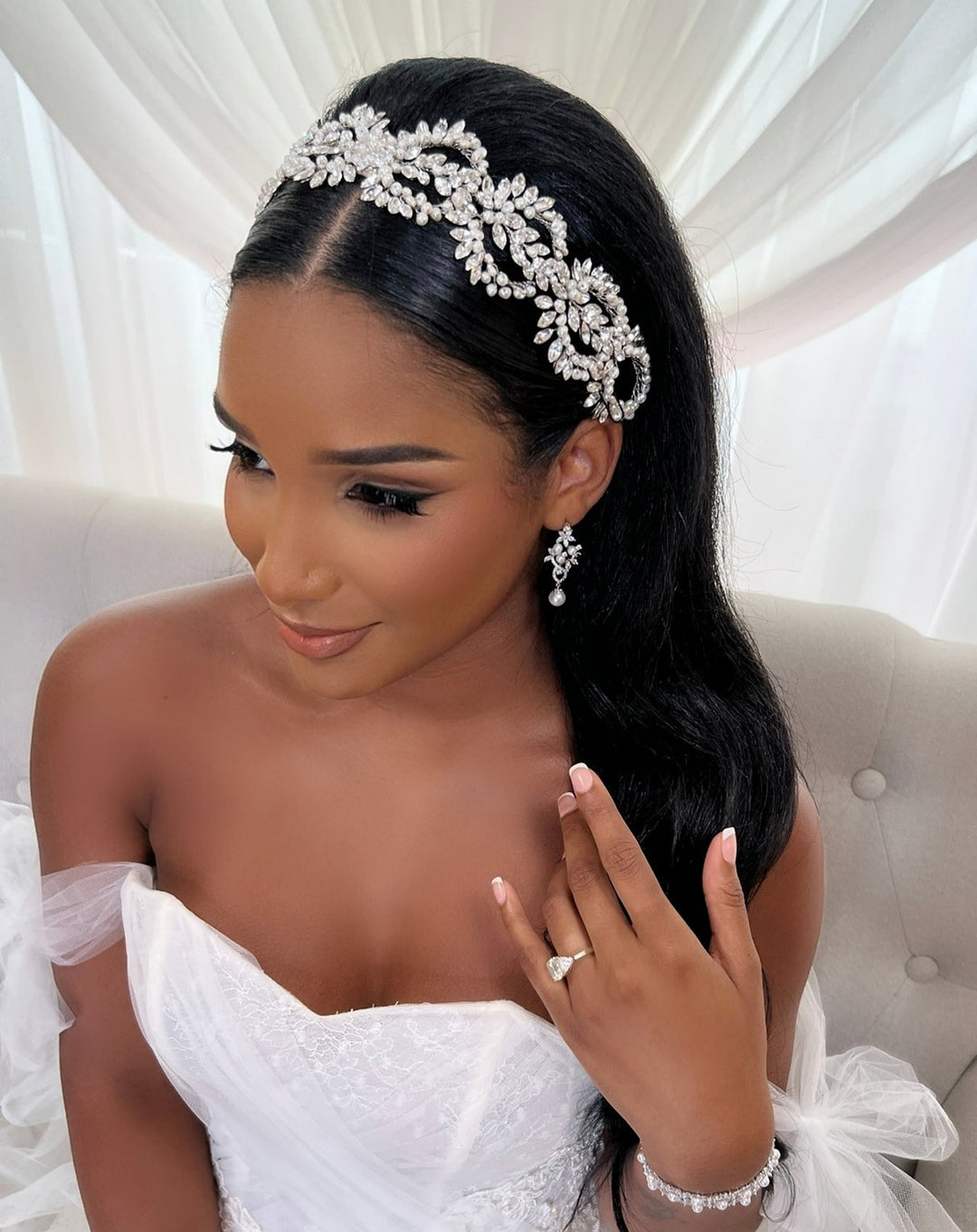 A bride with long dark hair wears the Crystal Wreath Hair Vine headband, diamond earrings, and a strapless white gown. Seated on a light sofa against sheer curtains, she touches her hair to display a ring and bracelet.