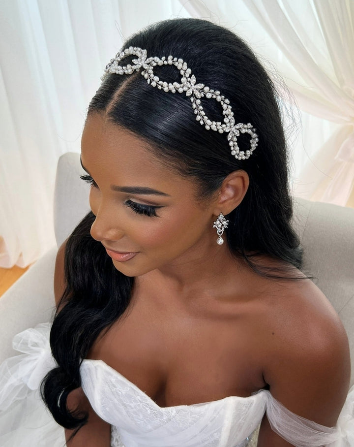A woman with long black hair wears Classy Pearl and Flower Looping Vine and matching pearl wedding earrings. She is dressed in a white, off-the-shoulder gown and sits against a light fabric backdrop, looking slightly to the side.