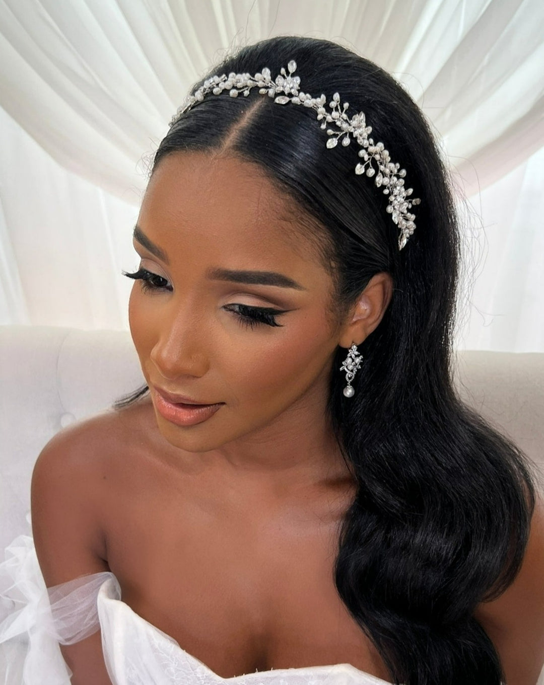A woman with long dark hair and striking makeup wears the Sweet Pearl and Swarovski Crystal Wedding Hair Vine (style no. 242719) and matching earrings. Shes in a white off-the-shoulder gown, seated against a white draped background, gazing down softly.
