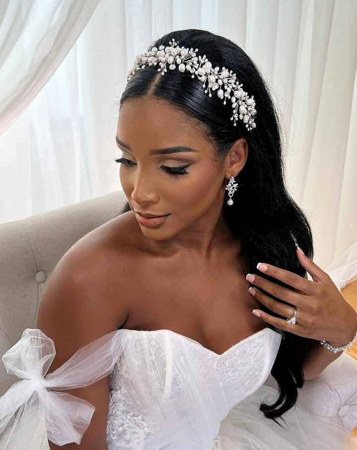 The bride sits gracefully in a white gown with a lace bodice, her look complemented by the Swarovski bridal tiara and matching earrings. Her long, dark hair cascades elegantly as she gently touches it, showcasing her engagement ring with poise. She is  wearing a Cluster Pearl and Crystal Bridal Vine (style no. 242704).