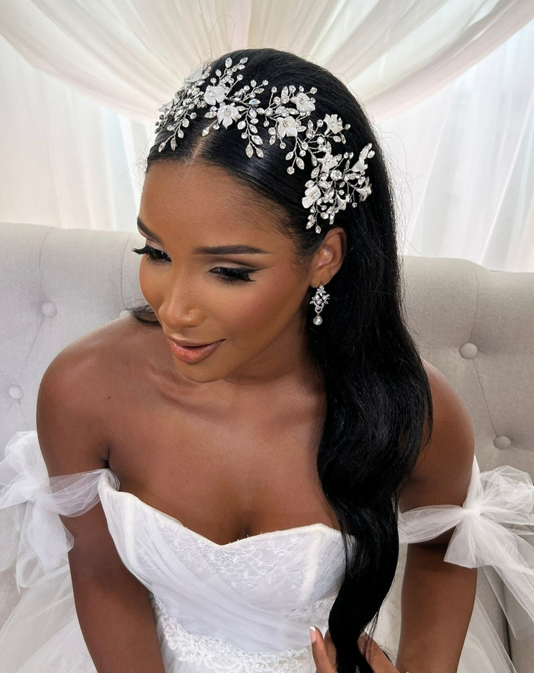 A bride with long dark hair, adorned with the Swarovski Crystal and Flower Branch Hair Vine (style no. 242736) and matching earrings, sits on a light-colored sofa in an off-shoulder white wedding gown, posing against draped fabric.