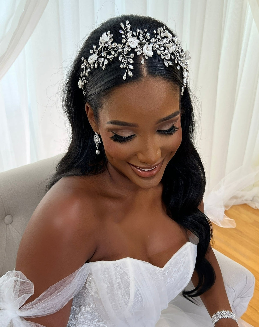The smiling bride wears an off-the-shoulder white gown and the Swarovski Crystal and Flower Branch Bridal Hair Vine (style no. 242736) hair combs. Her long, dark hair cascades elegantly over her shoulders, matching her floral earrings against sheer white curtains in the backdrop.