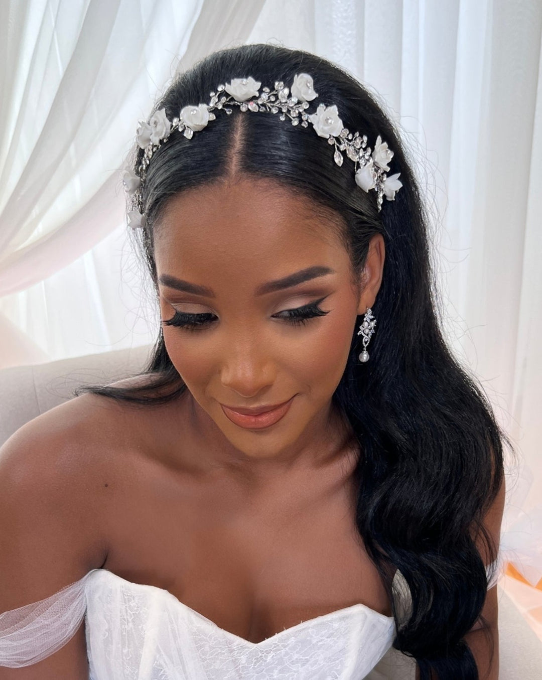 The bride with long black hair sits gracefully, adorned with a Swarovski wedding hair vine and pearl earrings. Her elegant white, off-the-shoulder gown is complemented by soft makeup featuring defined eyes and nude lips. Light curtains in the background add a serene touch. Product: Romantic Flower Vine, style 242720.