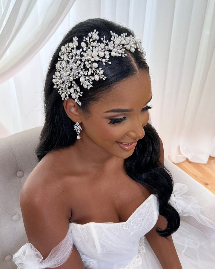 A bride dons a white strapless gown and the Dramatic Asymmetrical Pearl and Crystal Bridal Hair Vine (style no. 242703) in her long, soft waves of dark hair. She wears matching pearl drop earrings and sits on a light-colored chair against a softly draped white fabric background.