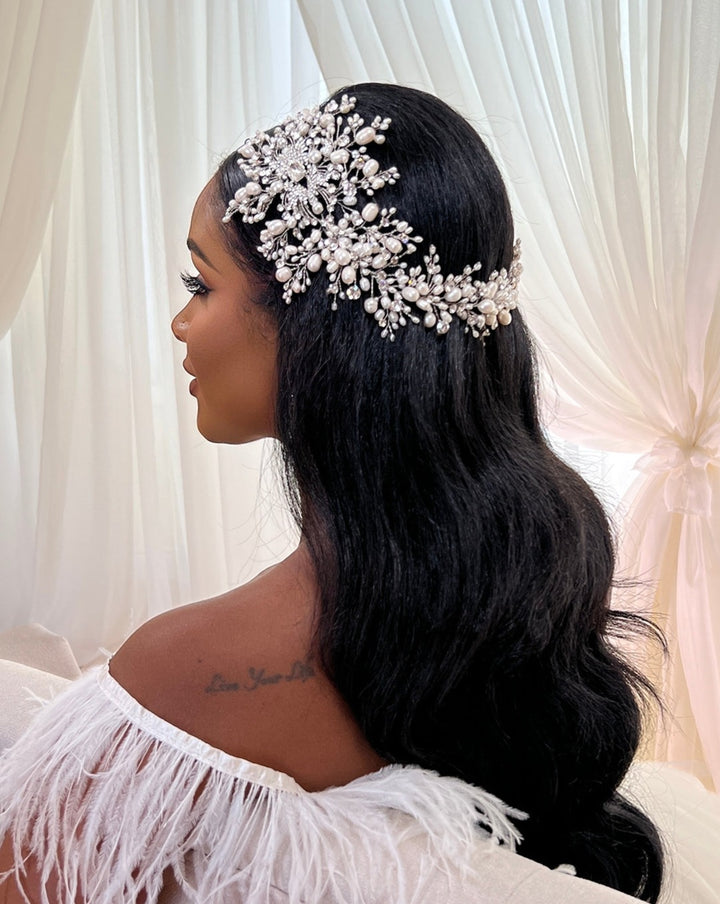 A woman with long, wavy black hair sits wearing a Swarovski bridal headpiece called Dramatic Asymmetrical Pearl and Crystal Vine style no. 242703, extending gracefully from her head to one side. Her off-the-shoulder white dress with feathered details is set against soft, white curtains.