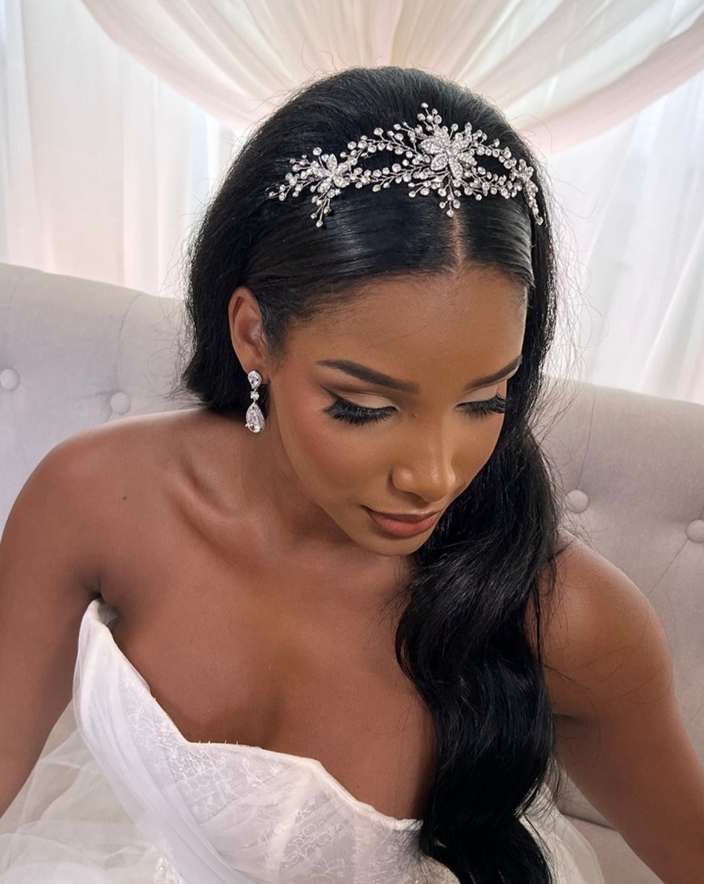 A woman in a strapless white dress sits on a light gray couch, her long dark hair adorned with a Swarovski crystal hair vine. She wears dangling CZ bridal earrings, and sheer white draped curtains frame the background.