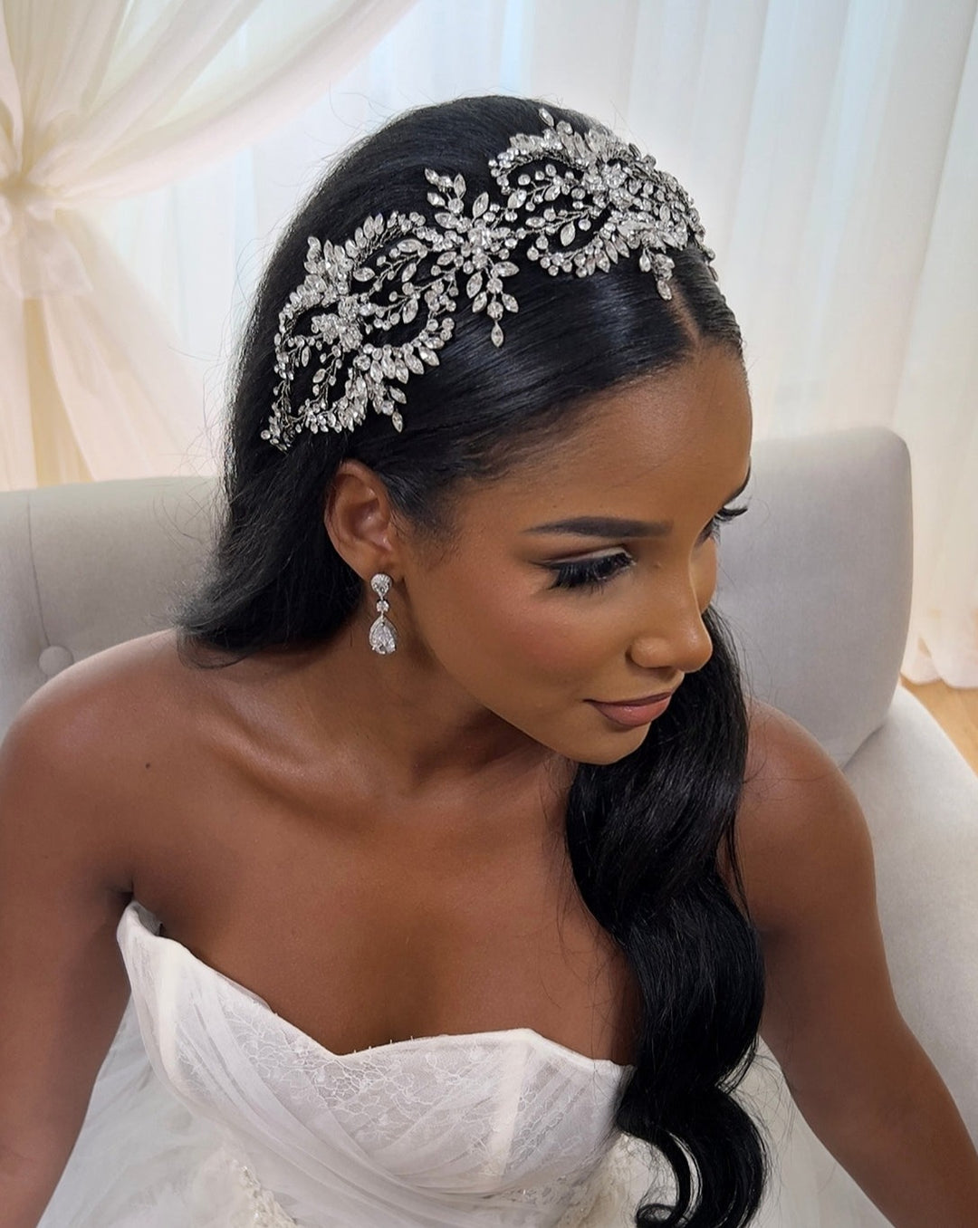 A woman in a strapless white gown sits on a light gray couch, wearing the sparkling Dramatic Wreath Vine Style no. 242682 headband and matching drop earrings. Her long black hair cascades over her shoulder, while sheer white curtains in the background enhance the ethereal ambiance.