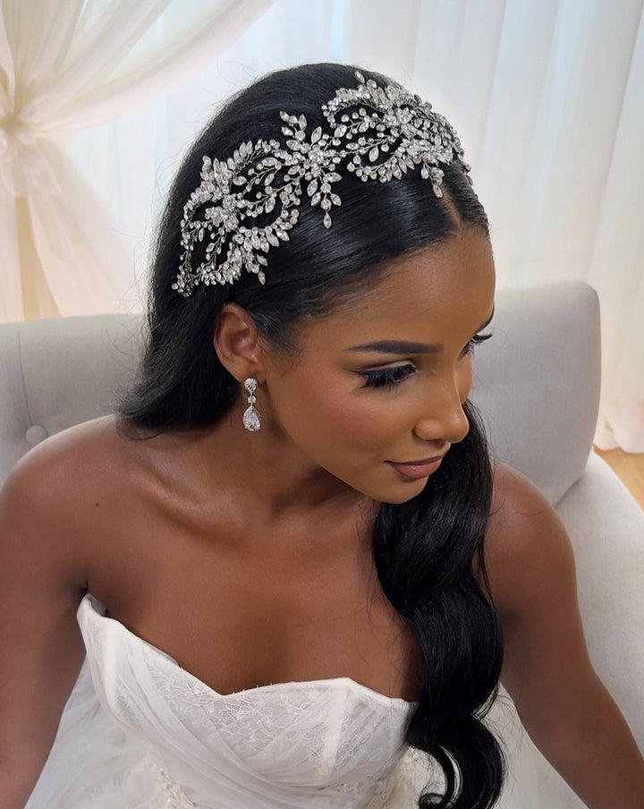 A woman in a strapless white gown sits on a light gray couch, wearing the sparkling Dramatic Wreath Vine Style no. 242682 headband and matching drop earrings. Her long black hair cascades over her shoulder, while sheer white curtains in the background enhance the ethereal ambiance.