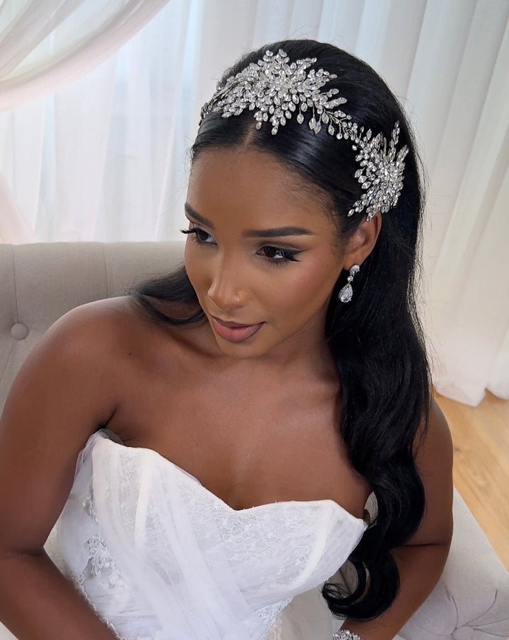 A woman in a strapless white bridal gown sits on a light chair. She has long, dark hair adorned with the Statement Crystal Vine, Style no. 242551, a Swarovski  crystal wedding headpiece, and wears drop earrings. Her makeup is elegant with sheer curtains visible in the background.