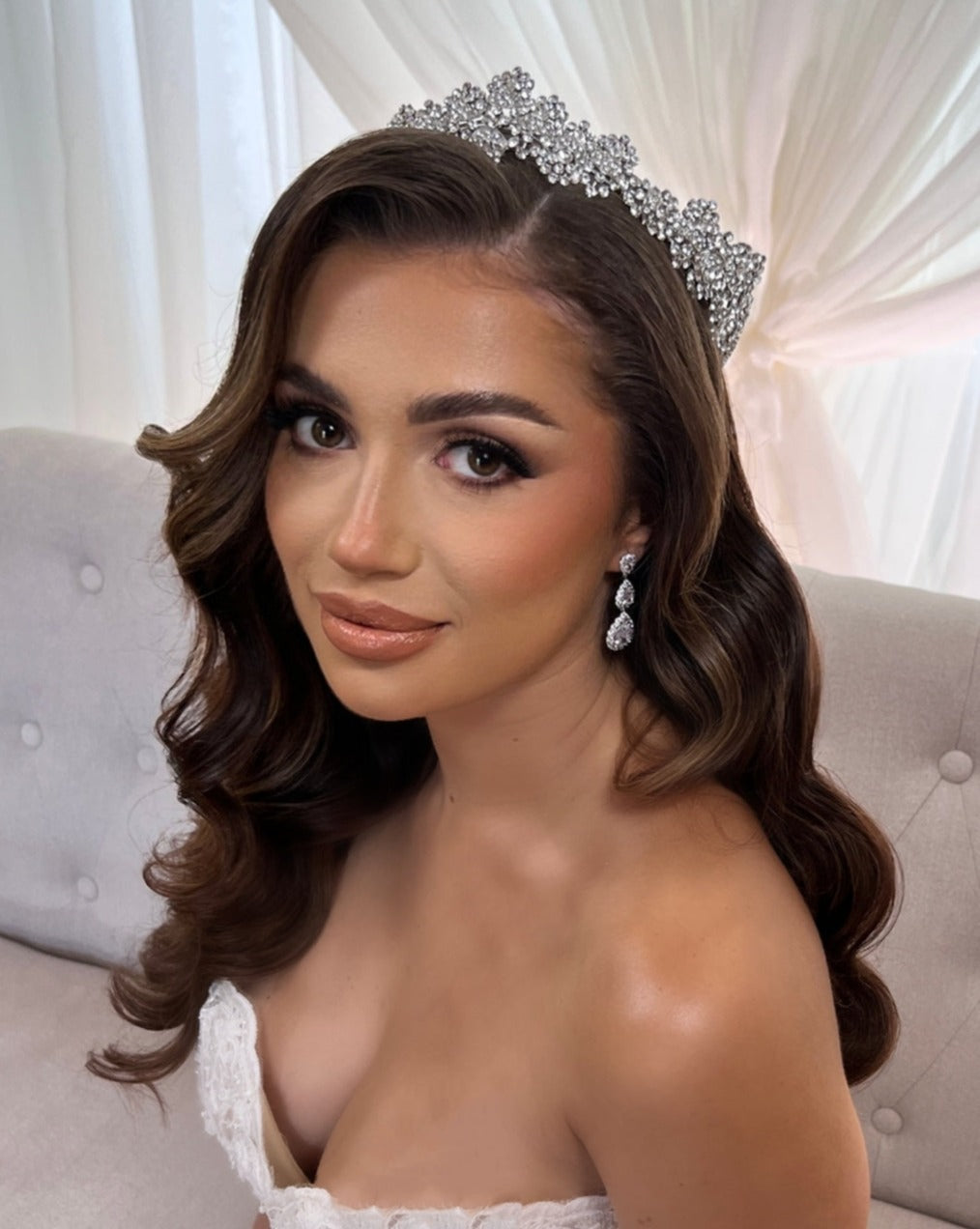 A woman with long, wavy brown hair wears a Crystal Wedding Tiara (style no. 80039) and sparkling drop wedding earrings. She is in a strapless white gown, seated on a light gray tufted couch against sheer white curtains.