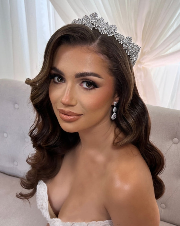 A woman with long, wavy brown hair wears a Crystal Wedding Tiara (style no. 80039) and sparkling drop wedding earrings. She is in a strapless white gown, seated on a light gray tufted couch against sheer white curtains.