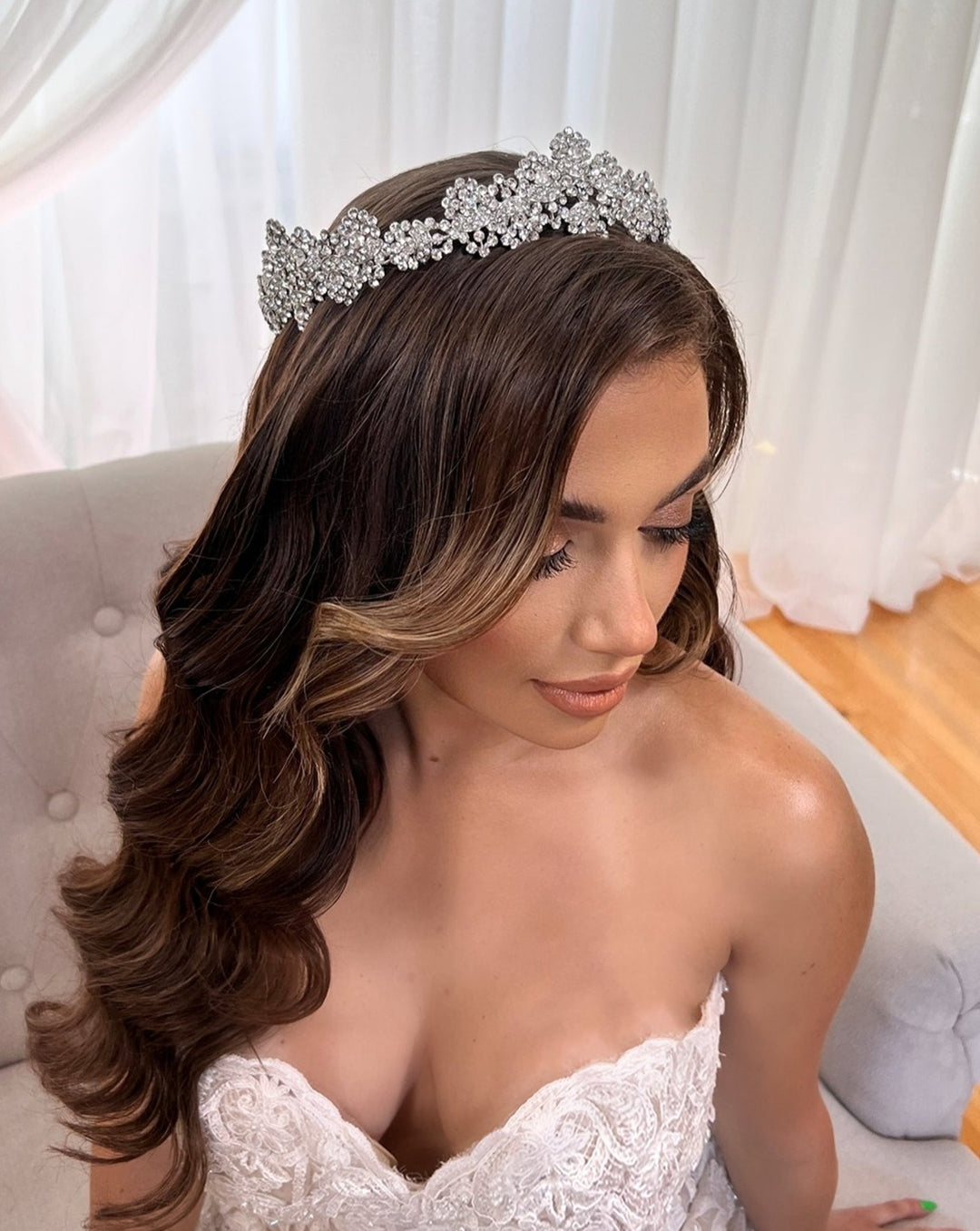 A woman with long, wavy hair adorned by a Swarovski Crystal Wedding Tiara style no. 80039 sits on a light gray sofa. Her off-shoulder white lace dress shimmers against a backdrop of soft, white curtains and a wooden floor, completing the enchanting scene.